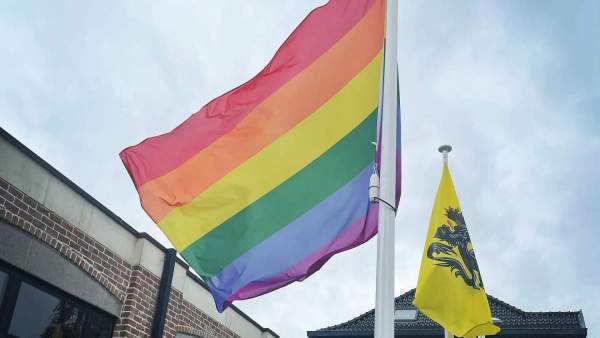 Een regenboogvlag aan een vlaggenstok die lichtjes wappert in de wind, naast drie andere vlaggenstokken meer op de achtergrond met de Vlaamse vlag en de vlaggen daarachter zijn moeilijk zichtbaar. De vlaggen staan aan het gemeentehuis van Kortenaken.