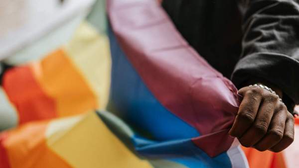 Een regenboogvlag die wordt vastgehouden door een person van wie enkel de linkerarm in beeld is.