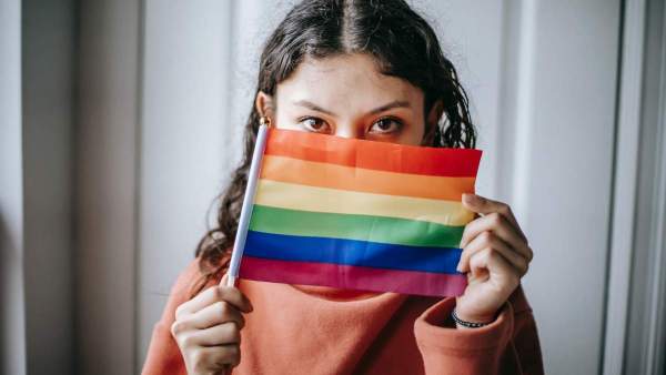 Een persoon houdt een regenboogvlag voor hun gezicht, enkel de ogen zijn zichtbaar.