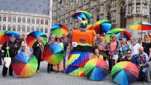 Groepsfoto op de Grote Markt van Leuven met Reus Iris van het Holebihuis. Vele mensen hebben een regenboogparaplu vast of een rond bordje in regenboogkleuren.