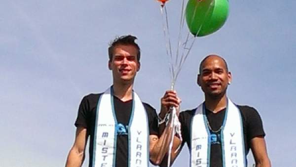 Een foto van twee personen die naast elkaar staan en samen een aantal gekleurde ballonnen van het Holebihuis in hun samengevouwen handen vasthouden. Ze dragen allebei een zwart T-shirt en een witte sjerp waarop staat gedrukt 'Mister Gay Vlaanderen'.