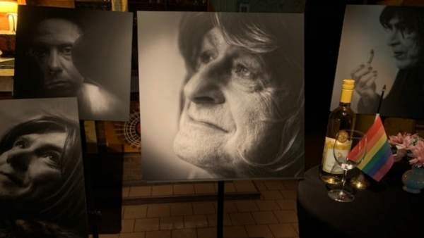 Enkele zwart-wit foto's van transgender personen op staanders in café Rocco, belicht door enkele spots. In het midden van het beeld is een zwart-wit close-up van Ghislaine Bonetto, een vrouw met lichtgekleurde haren die naar links kijkt en lacht. Rechts van de foto van Ghislaine staat een tafel met zwart tafelkleed waar een leeg wijnglas, fles alcohol en een regenboogvlaggetje.