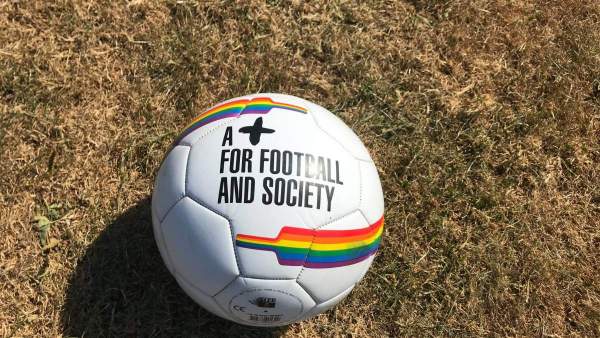 Een witte voetbal op het gras, er staan regenbogen op en in zwarte letters 'A + for football and society'