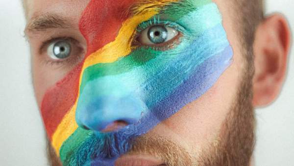 Close-up van een persoon met kort blond haar en een blonde baard die een schuine regenboog op het gezicht heeft geschilderd