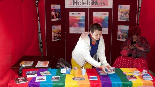 Sfeerfoto van de binnenkant van de infostand van het Holebihuis, op een tafel ligt een regenboogvlag en verschillende folders, een persoon in een witte cardigan legt folders op tafel en links in de achtergrond zit een drag queen in roze kledij op een stoel