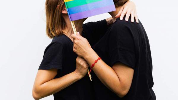 Twee personen in zwarte T-shirts die elkaar een kus geven, hun gezichten verscholen achter een regenboogvlagje dat ze in hun handen hebben