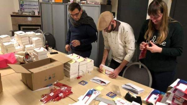 Een foto van drie vrijwilligers van de werkgroep Take Care, staan langs elkaar aan een tafel. De tafel ligt vol met brochures, dozen, lintjes, enveloppes, en dergelijke. De drie personen zijn aan het werk.