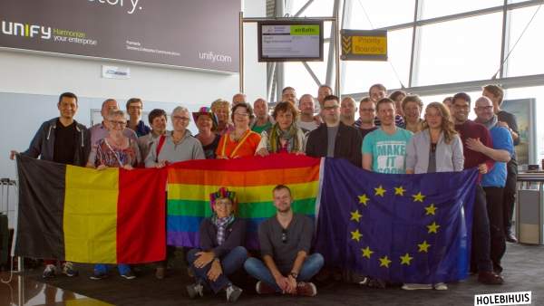 Een groepsfoto van de vrijwilligers van het Holebihuis in een vliegveld, ze houden omhoog een Belgische vlag, regenboogvlag en Europese vlag